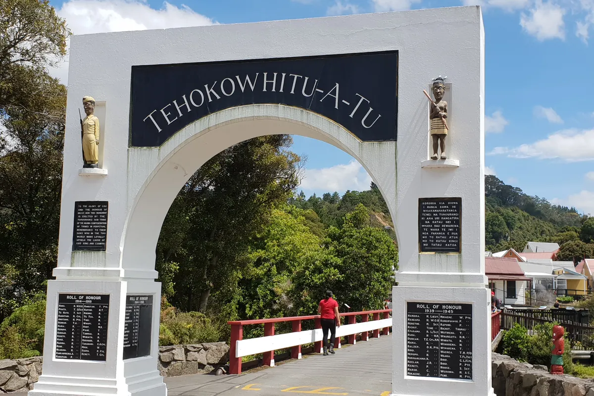 Den vita minnesporten vid ingången till Whakarewarewa by, med en gångbro och ett rött tak över ett hus bortom